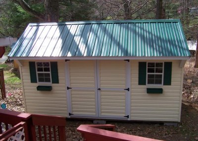 A 10x16 vinyl shed with a metal, carriage style roof. Shed has two windows with green shutters and flower boxes. Shed has a set of double doors