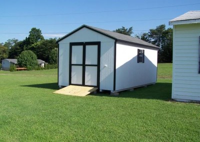 A white painted shed with a black shingled, a-roof style roof. Shed has black trim, a solid double door, two windows with black shutters, and a treated wooden plank