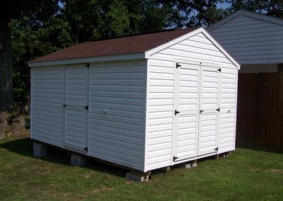 A white vinyl shed with a shingled a-roof style roof and white trim. shed has a double door on gable end and a 3 foot vinyl door on side