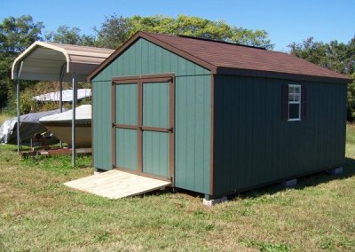 A 12x16 sized green shed with brown shingles and trim. Shed has a-roof style roof, a set of double doors, two windows with shutters, and a treated wooden ramp