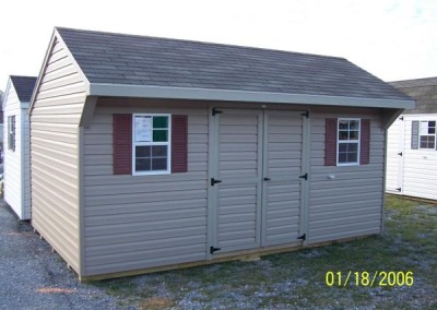A vinyl 10x16 shed with a shingled carriage style roof. Shed has a set of double doors and two windows with shutters