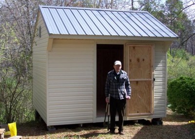 A white vinyl, 12x12 shed with a metal carriage style roof. Shed has a gable vent and one 3 foot wide door
