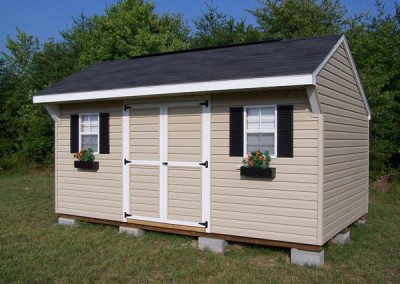 A vinyl 10x16 shed with a shingled a-roof style roof. Shed has a shingled carriage style roof and two windows with black shutters and flowerboxes. Shed has a double door