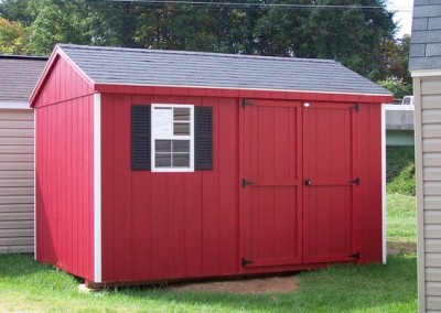 A red painted 8x12 shed with a shingled roof. Shed has one window with shutters and a solid double door.