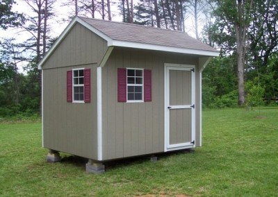 An 8x10 painted shed with a shingled carriage style roof. Shed has a single painted door and two windows with shutters