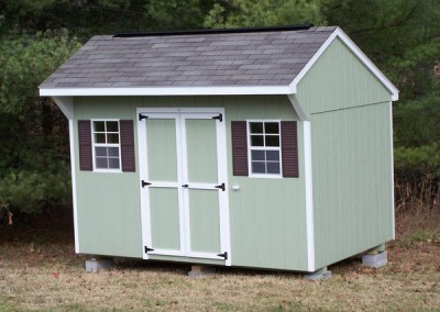 A light green painted shed with a shingled, carriage style roof. Shed has a set of double doors and two windows.