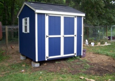 A blue painted 8x8 shed with white trim. Shed has a set of double doors and a window with white shutters. The roof is a-roof style and is shingled.