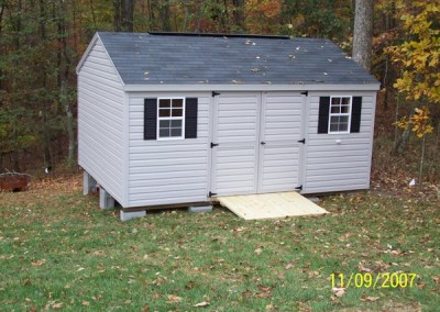A 12x16 vinyl shed with a set of double doors and two windows with shutters. Shed has a treated wooden ramp