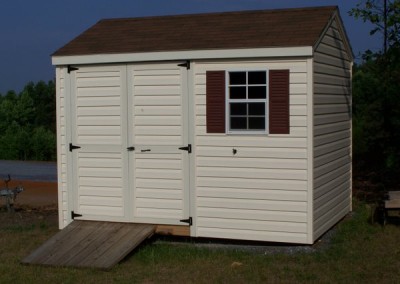 A 8x10 vinyl shed with a shingled, a-roof style roof. Shed has a set of double doors, a window with shutters, and a wooden ramp